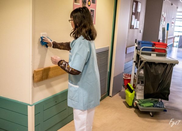 Une femme agent d'entretien en train de faire le ménage dans un couloir de centre hospitalier ou de ce type.