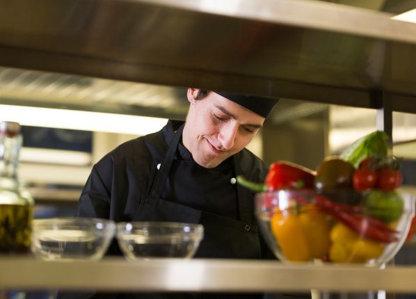 Un cuisinier derrière des étagère dans une cuisine de restaurant en train de sourire et de composer un plat